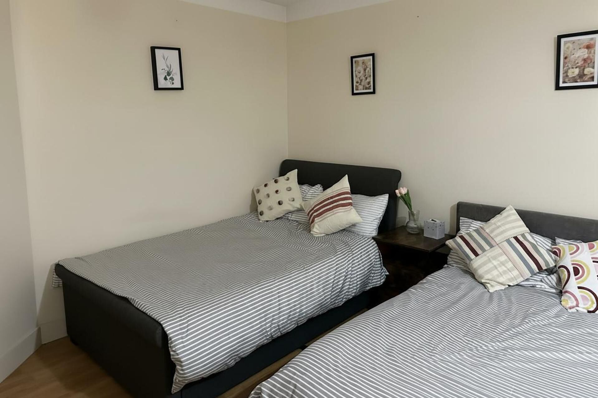 A bedroom with two single beds, each with striped bedding and multiple pillows. There are framed pictures on the walls and a small table with a vase and a tissue box.