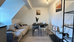 A modern living room with a sofa, dining table, and chairs. There is a ceiling light, a framed picture on the wall, and a plant on the table. - Oak Wembley Stadium Serviced Apartment by Edens (Wembley)