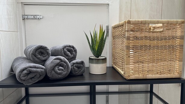 A bathroom with a white door, a black metal shelf holding rolled towels, a potted aloe vera plant, and a woven basket. - Oak Wembley Stadium Serviced Apartment by Edens (Wembley)