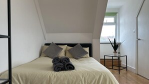 A bedroom with a bed, pillows, a small table with a plant, and a window. - Oak Wembley Stadium Serviced Apartment by Edens (Wembley)