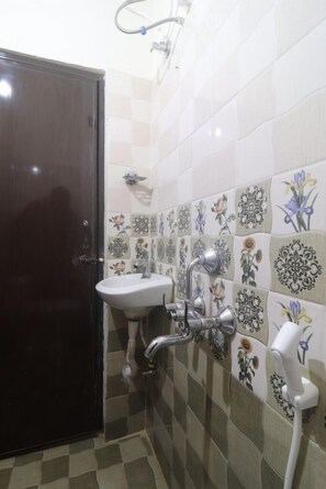 Une salle de bain avec une porte sombre, un lavabo blanc et une douche équipée d’un pommeau de douche à main. Les murs sont recouverts de carreaux aux motifs floraux.