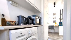 Een kleine keuken met een witte wasmachine, waterkoker en magnetron op het aanrecht.