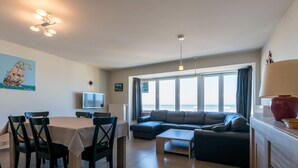 A modern living room with a dining area, large windows, and a television. - Apartment in Koksijde for 8 Person (Koksijde)