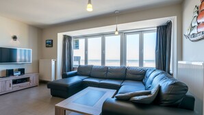 A modern living room with a sectional sofa, a flat-screen TV, and a large window with a view of the beach. - Apartment in Koksijde for 8 Person (Koksijde)