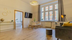 Un salon moderne avec un canapé, un coin repas avec des chaises, un téléviseur fixé au mur et une grande fenêtre donnant sur des immeubles.