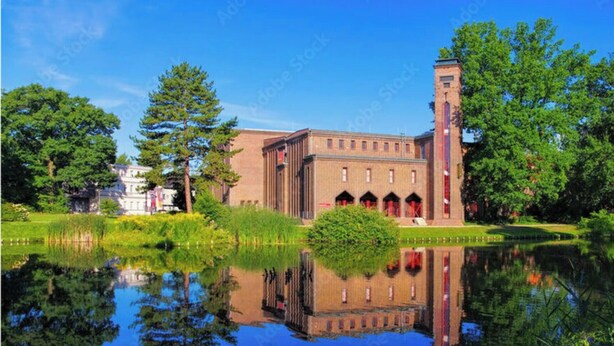 Un bâtiment historique doté d’une tour centrale, entouré d’une végétation luxuriante et bordé d’une étendue d’eau calme dans laquelle se reflète la structure.