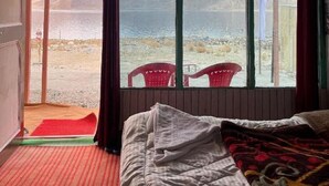 A room with a bed, a window with a view of mountains and a body of water, and a red chair outside. - Padma Cottage Pangong (Leh)