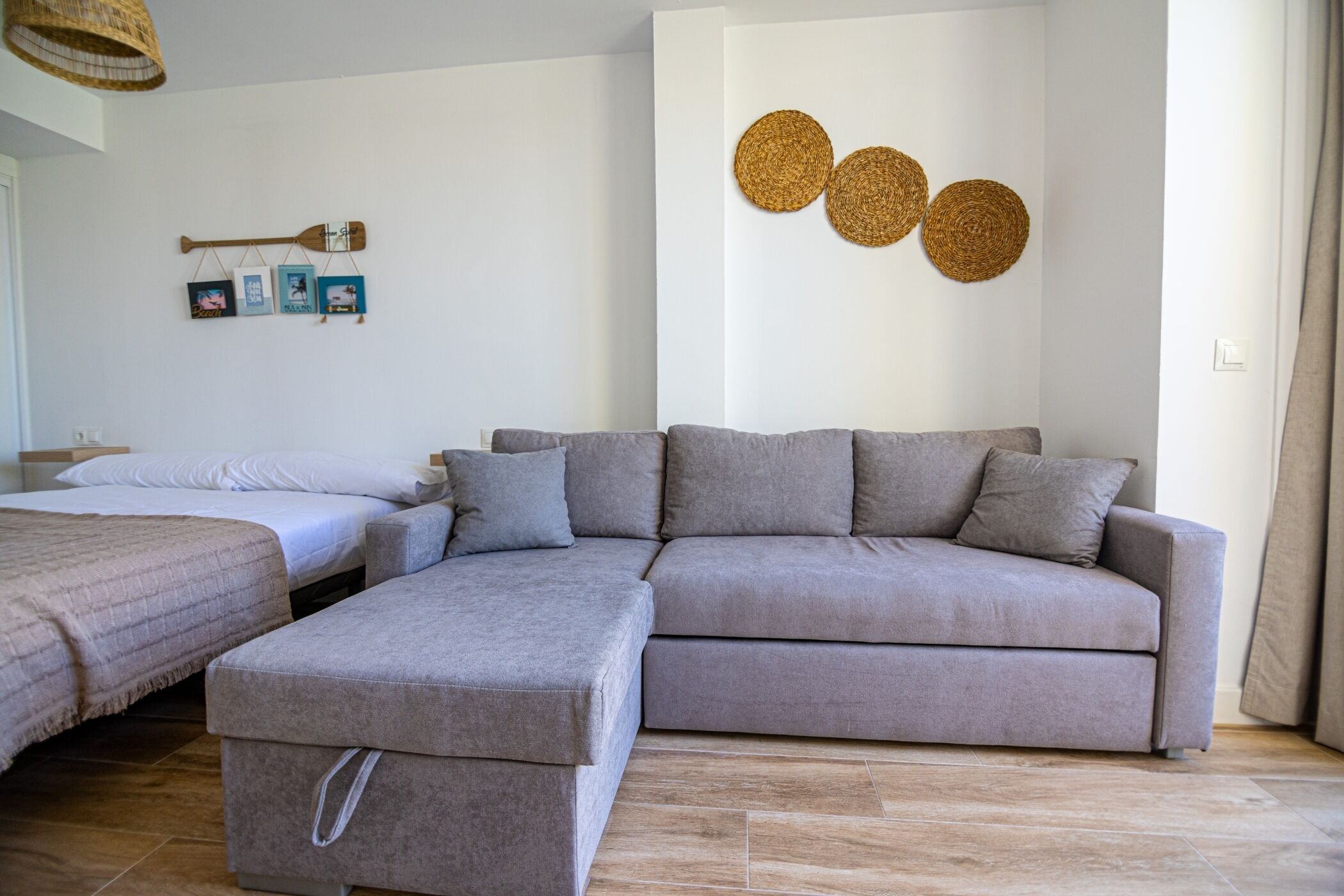 A modern living room with a grey sectional sofa, a matching ottoman, and a small coffee table. There are two beds with white linens and a wall-mounted shelf with decorative items.