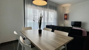Ein modernes Hotelzimmer mit Esstisch, Stühlen, einer Sitzecke, einem Fernseher und einem großen Fenster mit durchsichtigen Vorhängen.