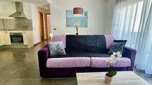 Un salon moderne avec un canapé violet, une petite table, une plante en pot et une cuisine en arrière-plan.