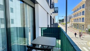 Un balcon avec une table et des chaises, donnant sur une rue animée par des piétons et bordée d’immeubles.