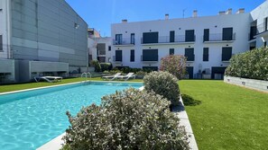 Uma piscina com espreguiçadeiras, um gramado bem cuidado e um edifício moderno com varandas.