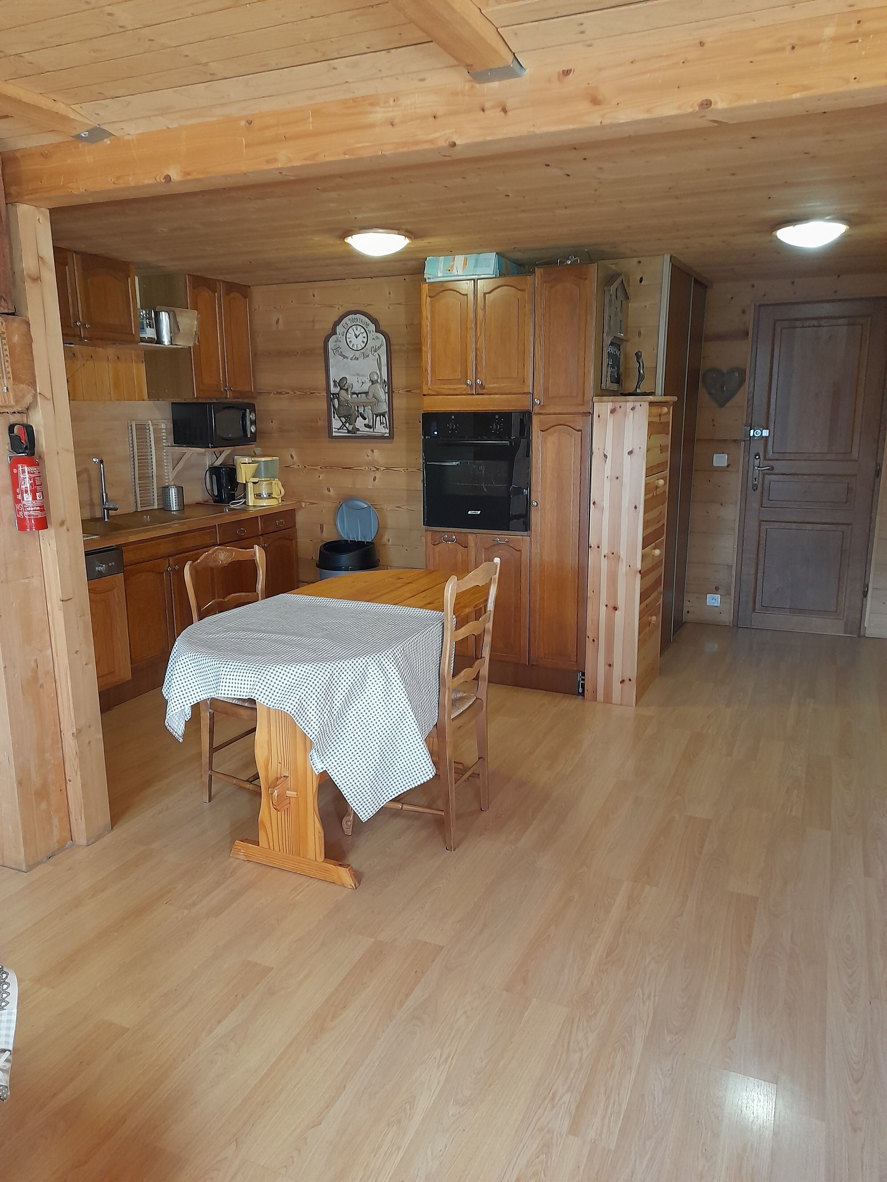 A wooden interior with a dining table, chairs, a kitchen area including a microwave and oven, and a wooden cabinet.