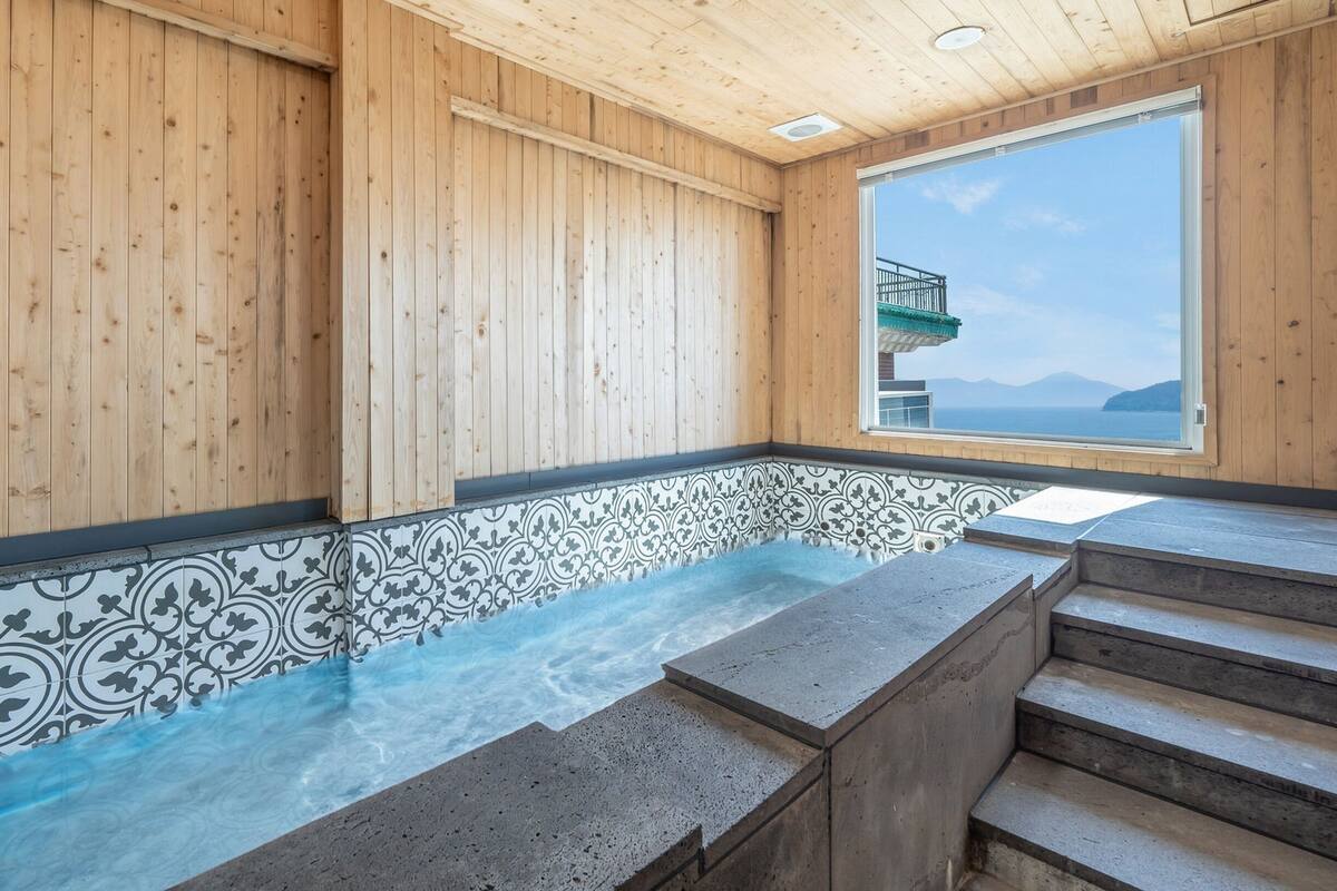 A wooden interior with a stone-tiled hot tub, a view of the sea, and a staircase leading to a balcony.