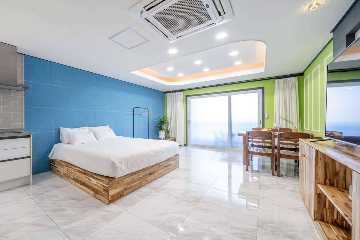 A modern hotel room with a large bed, a kitchenette, a dining area, and a sliding glass door to the outside.