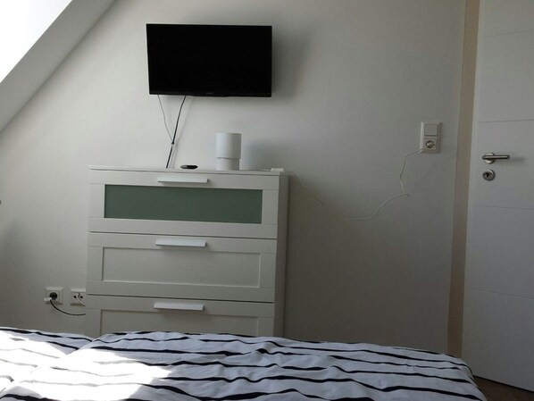 A bedroom with a bed, a white dresser, a flat-screen TV mounted on the wall, and a door with a handle.