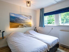 Une chambre à coucher avec un lit, un tableau représentant un coucher de soleil sur l’océan, une fenêtre donnant sur un paysage verdoyant et un radiateur.
