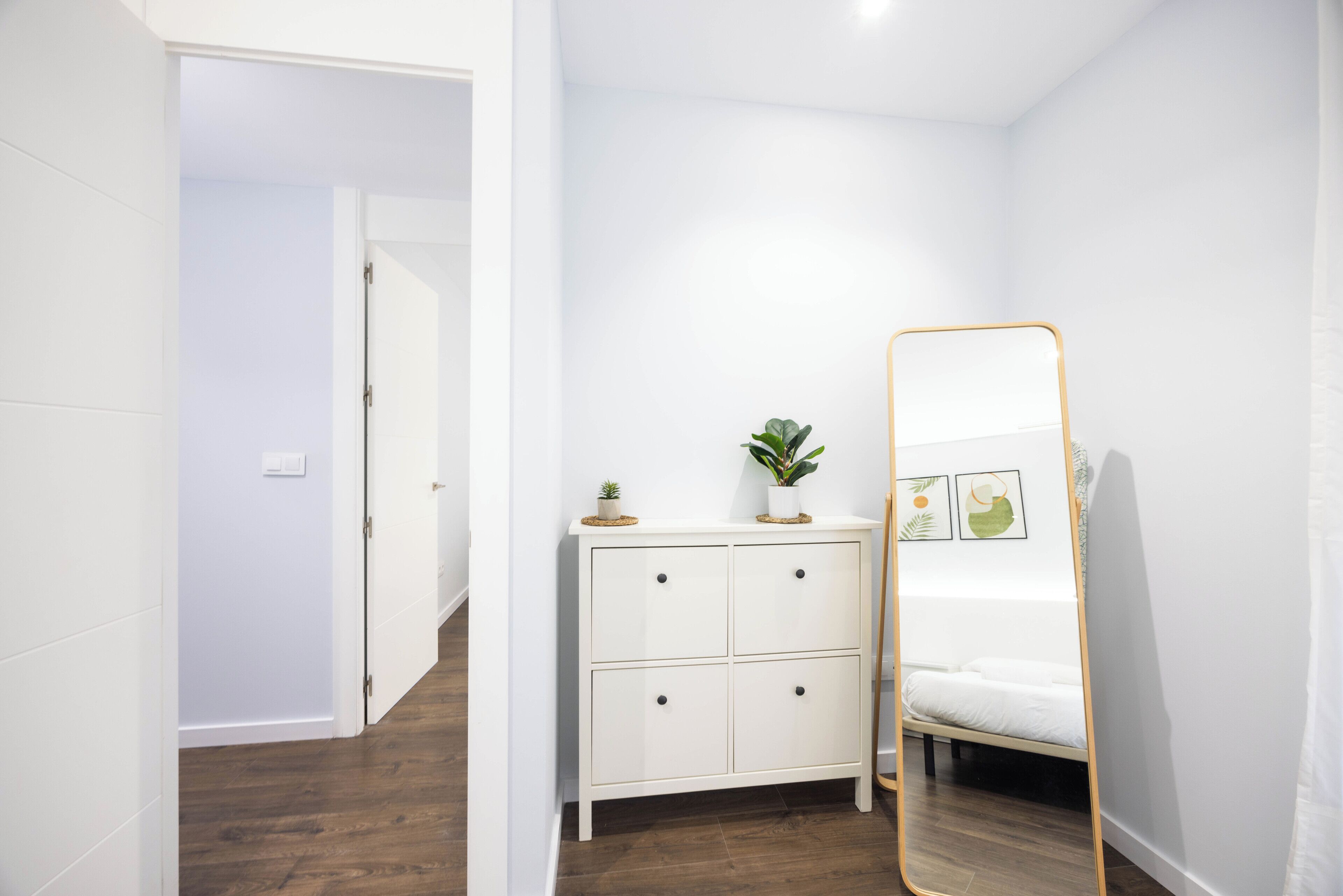 Un dormitorio con cama, una mesita de noche con planta, un espejo y una puerta que da a otra habitación.