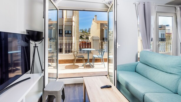 A modern living room with a sofa, a TV, a small table, and a balcony with a table and chairs. - Apartamentos Turísticos Metrópolis (Cartagena)