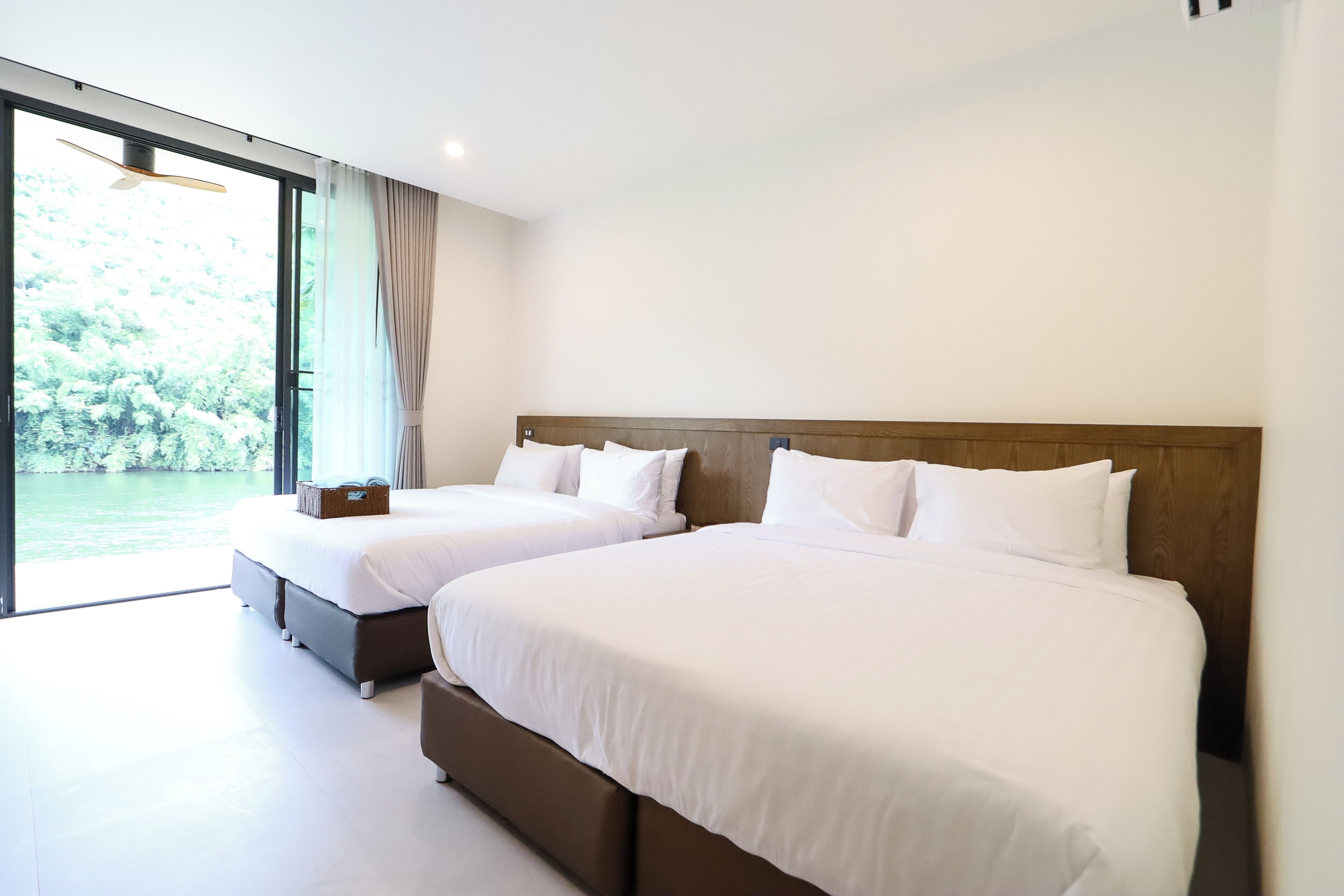 A modern hotel room with two beds, a large window with a view of greenery, and a wooden headboard.