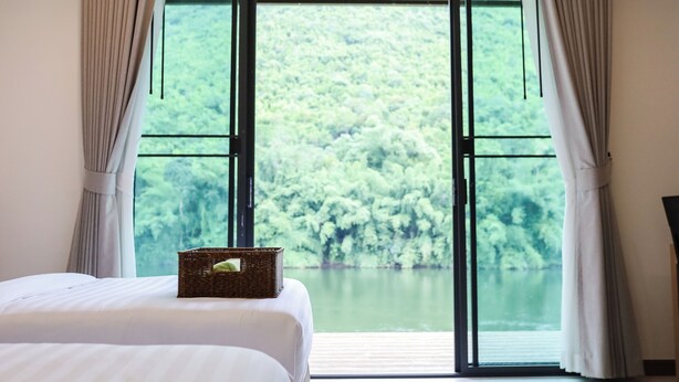A bedroom with a large window, a bed, a basket on the bed, and a ceiling fan. - Tiger House River Kwai (Si Sawat)