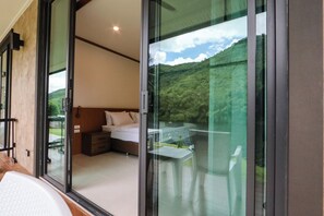 A bedroom with a bed, a nightstand, and a window overlooking a garden and a body of water. - Tiger House River Kwai (Si Sawat)