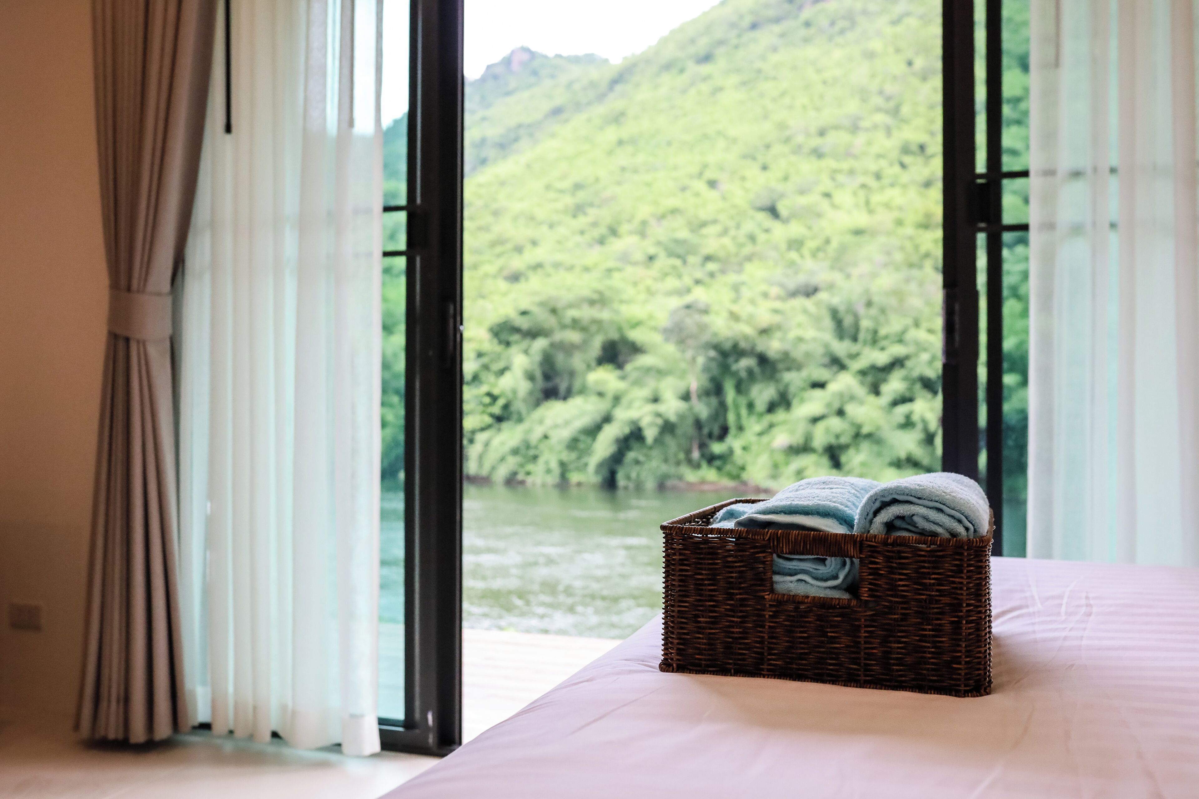 A bedroom with a large window overlooking a river and forest. A wicker basket with towels is on the bed.