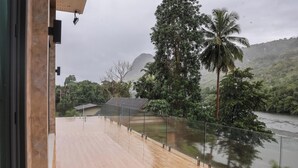 A wooden deck with a glass railing overlooking a river, surrounded by lush greenery and a mountain in the background. - Tiger House River Kwai (Si Sawat)