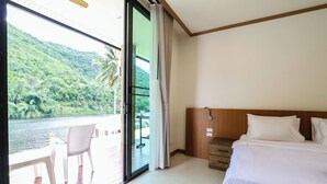 A hotel room with a large sliding glass door leading to a balcony with a view of a river and greenery. There is a bed with white bedding and a wooden headboard. - Tiger House River Kwai (Si Sawat)