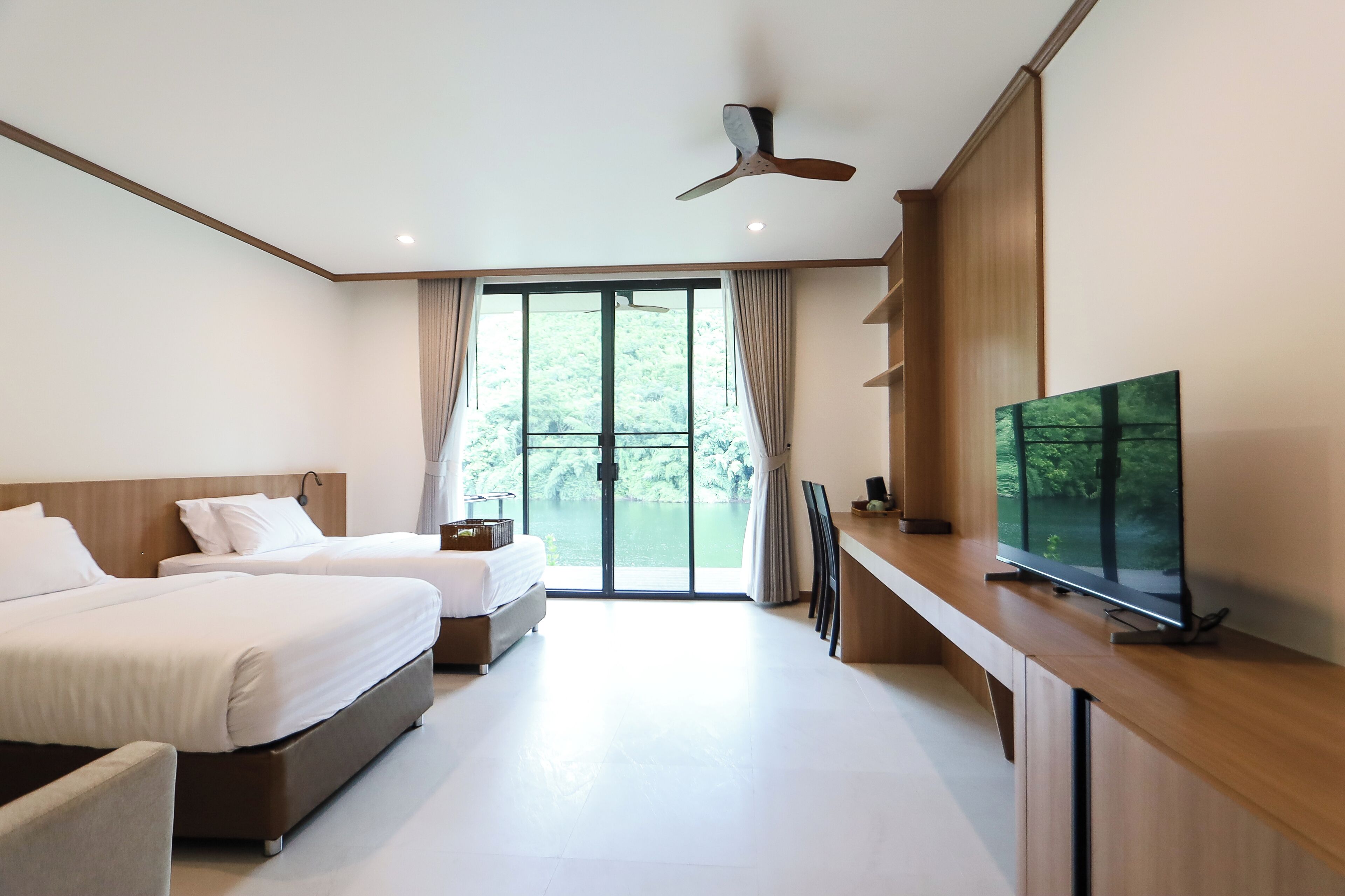Une chambre d’hôtel moderne dotée de deux lits, d’un téléviseur à écran plat, d’un ventilateur de plafond et d’une grande fenêtre donnant sur un paysage verdoyant.