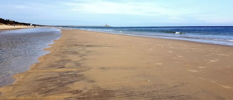 Una amplia playa arenosa con olas suaves, cielo despejado y un muelle distante.