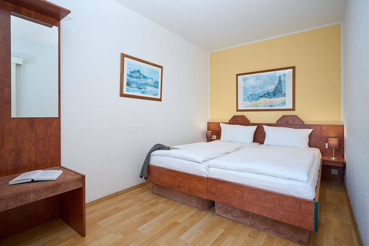 A hotel room with a wooden bed, a bedside table with a book, two framed pictures on the wall, and a wooden floor.