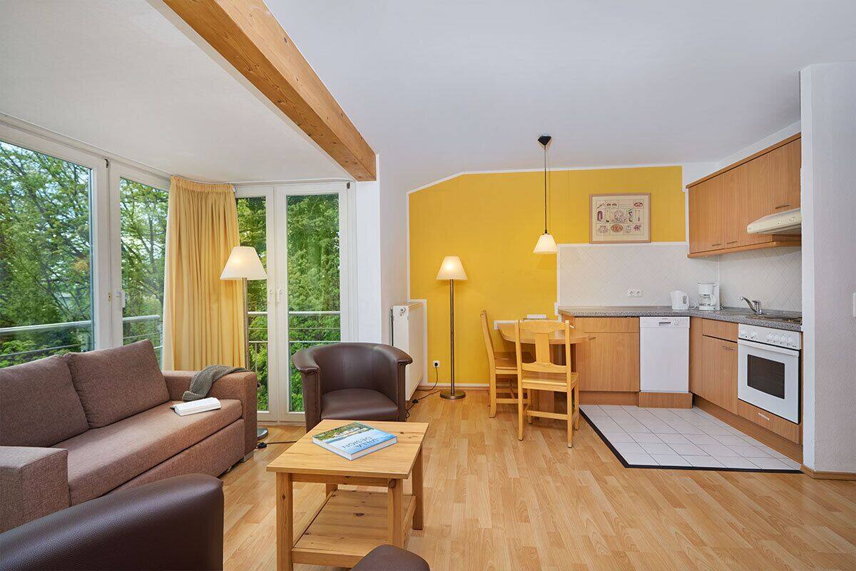 A modern living room with a sofa, armchair, and coffee table, a small kitchen with wooden cabinets and appliances, and large windows with curtains.