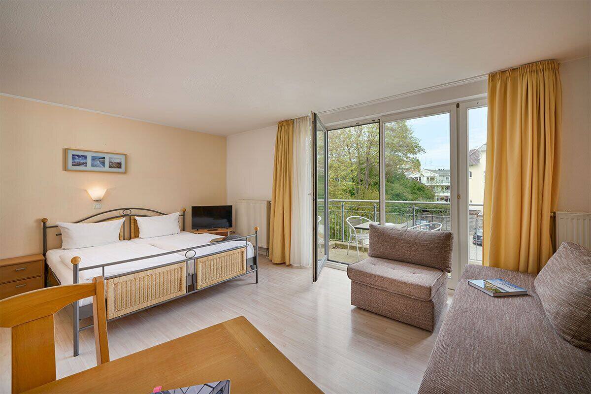A hotel room with a bed, a TV, a dining table, a sofa, and a balcony with a view of the outside.