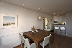 Een eethoek met een houten tafel, rieten stoelen, een hanglamp en een keuken op de achtergrond.