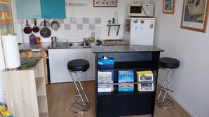 A compact kitchen with a microwave, sink, and two bar stools. There are books on a shelf and a framed picture on the wall. - Apartement de 1 à 4personnes 200m de la mer (La Turballe)