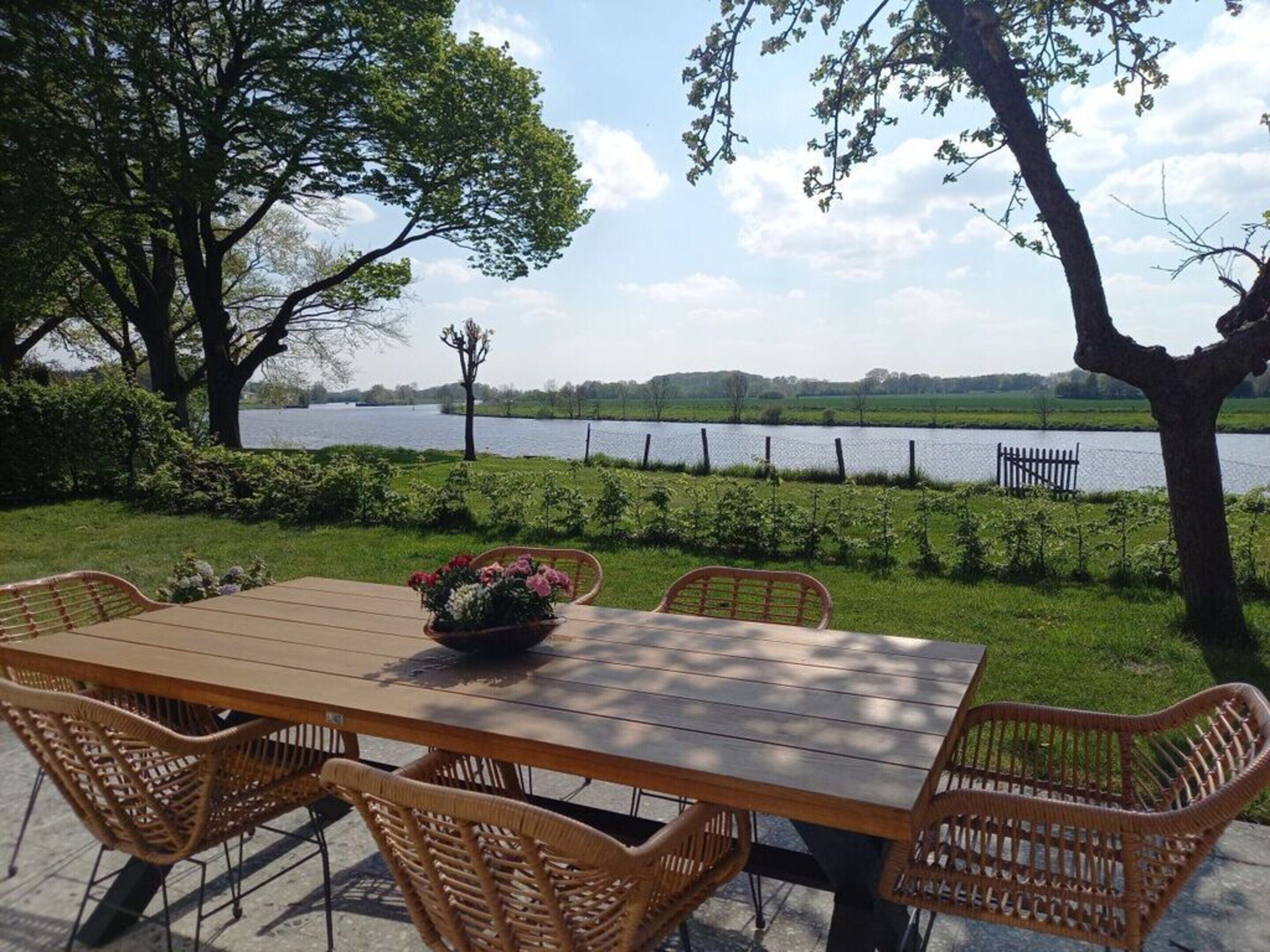 An outdoor dining area with a wooden table and wicker chairs, overlooking a lake with a fence and trees in the background.