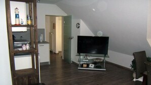 A living room with a flat-screen TV on a glass stand, a wooden bench, and a wicker chair. - Pommernweg 15 Merchant (Wangerland)