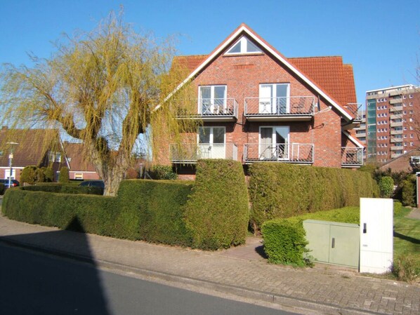 A two-story brick house with a sloped roof, a balcony on the second floor, and a well-maintained front yard with hedges. - Pommernweg 15 Merchant (Wangerland)