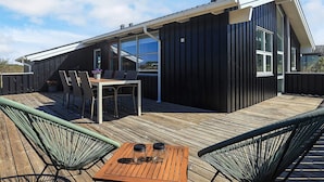 A modern house with a wooden deck, a dining area with chairs and a table, and a large chimney. - 4 Star Holiday Home in Hjorring (Hjørring)