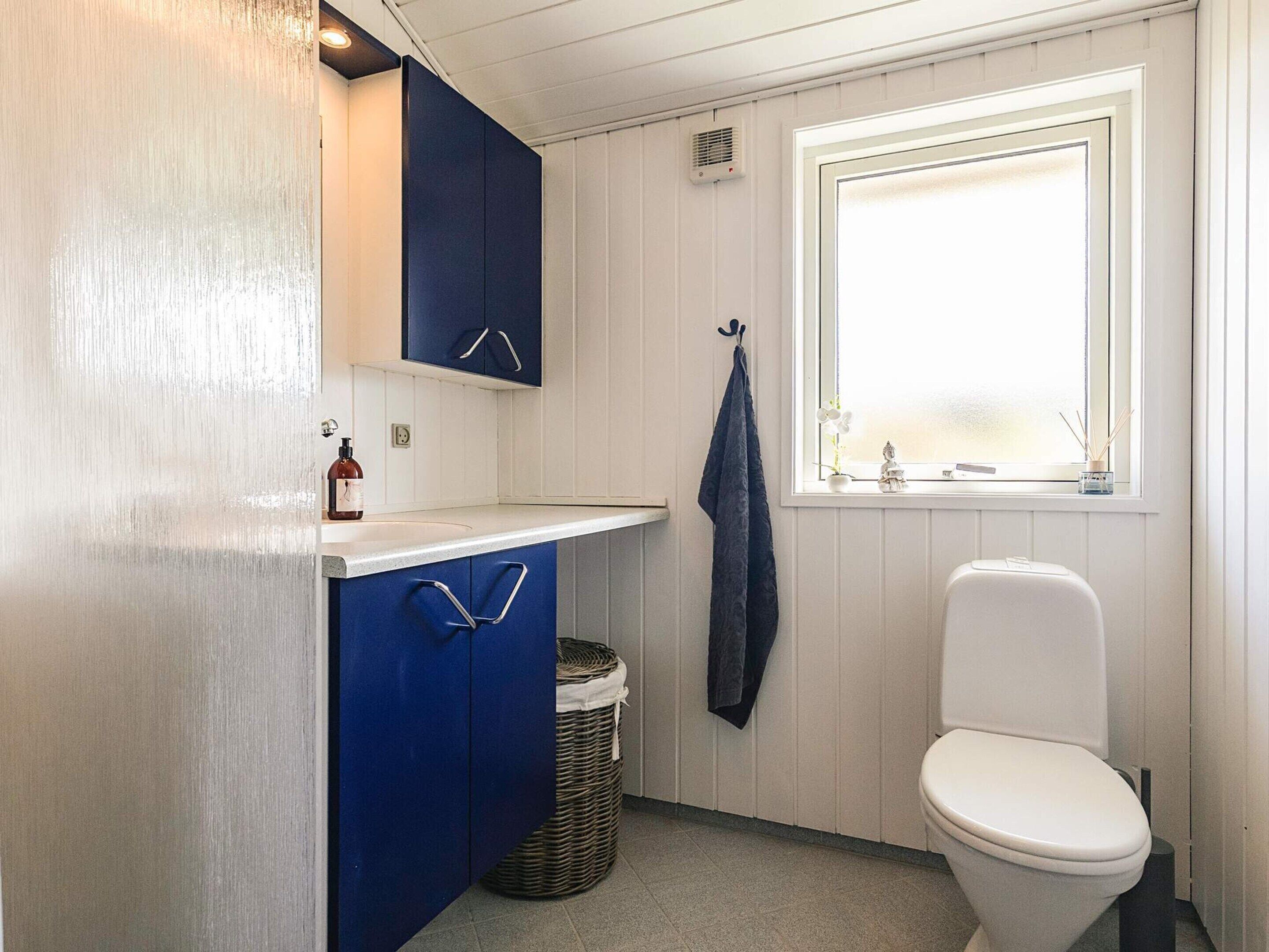 A compact bathroom with a white toilet, a sink with a blue cabinet, a wicker basket, and a window with a view.