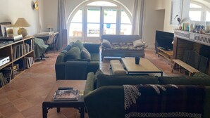 En stue med sofa, lænestole, sofabord og fjernsyn. Der er et bogskab med bøger og en lampe. Værelset har et stort buet vindue og et træloft.