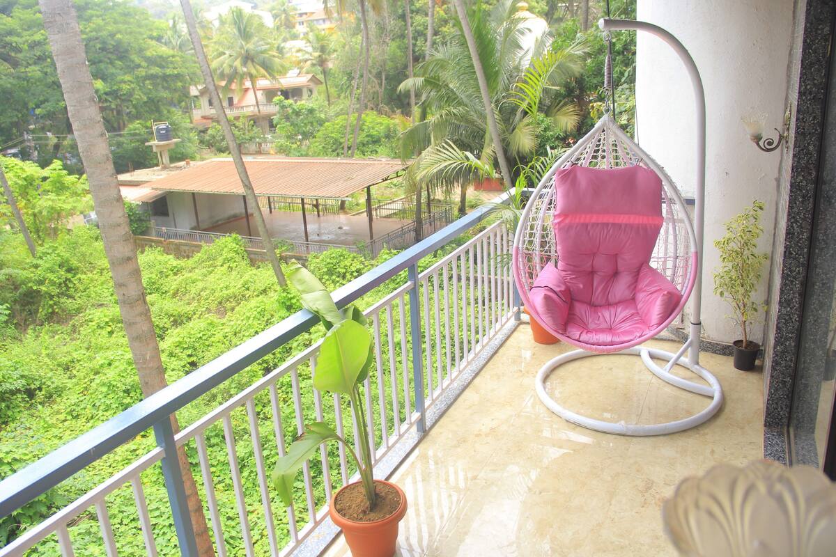 Un balcón con una silla colgante rosa, una planta en maceta y vistas de un paisaje verde exuberante con edificios al fondo.