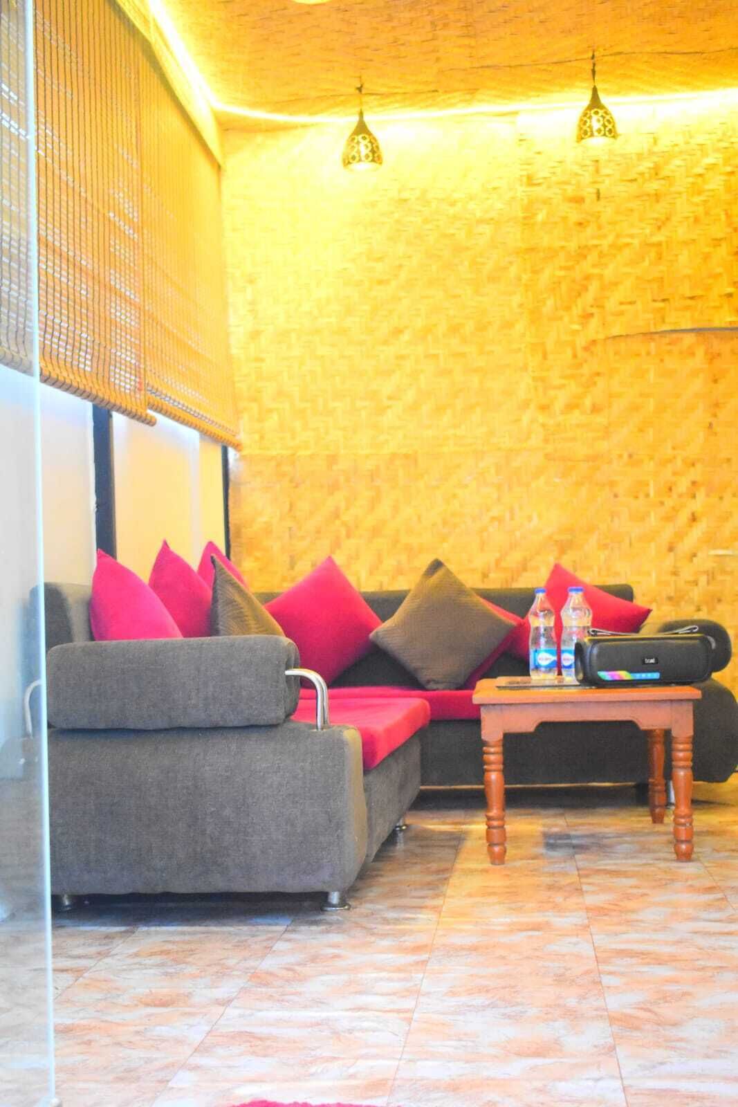 A cozy room with a grey sofa, red and brown cushions, a small wooden table with a water bottle and a bag, and two hanging lamps.