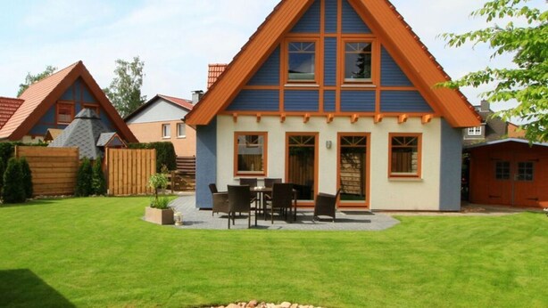 A house with a steeply pitched roof, a front porch with a table and chairs, and a well-maintained lawn. - Luerssen Lindenhopsweg (Wunstorf)