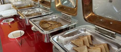 Een buffetopstelling met verschillende soorten eten in metalen bakken, waaronder belegde broodjes en koekjes, op een rode tafel met wegwerp borden en bestek.