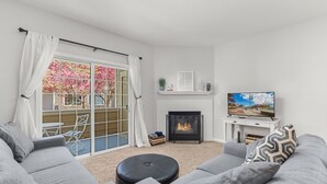 A modern living room with a fireplace, grey sofas, a television, and a view of cherry blossoms through a sliding glass door. - Beacon at the Lakes - Cozy & Bright 2bd/2ba Ground Level Condo (Windsor)