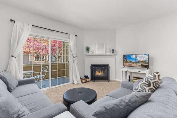 Un salon moderne avec une cheminée, des canapés gris, un téléviseur et une vue sur les fleurs de cerisier, visible à travers une porte-fenêtre coulissante.