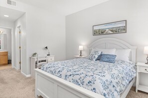 Une chambre avec un lit blanc, une table de chevet avec une lampe, une autre table de chevet avec une lampe, un tableau encadré au mur et une porte donnant sur une autre pièce.