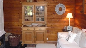 L’intérieur chaleureux d’un chalet présente un meuble en bois où sont exposés des verres, un panier en osier, un canapé blanc et une petite table avec une lampe.